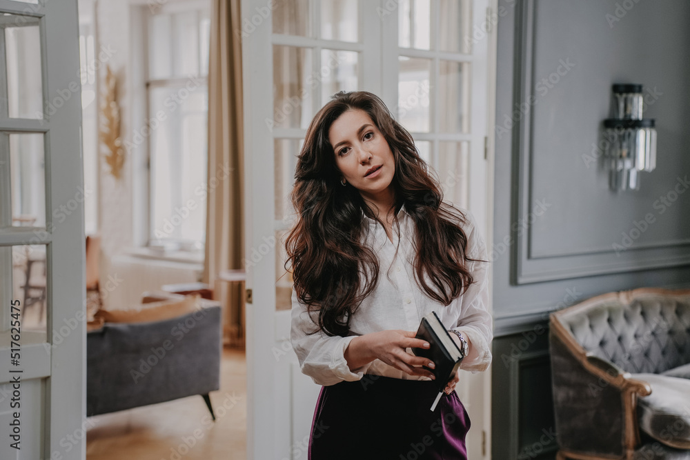 Calm brunette woman with wavy dark hair in white shirt and violet pants standing in living room, looking at camera, holding diary. Tired businesswoman at home after busy day at work. Business people.