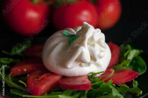 on a black plate is a salad of spinach and arugula greens, sliced tomatoes and Italian burrata cheese in close-up, a bunch of red tomatoes is visible in the background, side view.