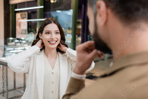 Positive woman wearing necklace near blurred boyfriend in jewelry shop.