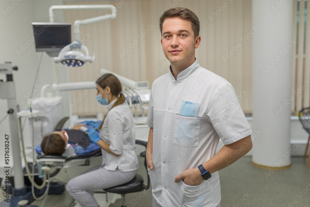 Obraz premium A male dentist looks at the camera against the background of a dental cabinet with a patient. Dental care and treatment concept