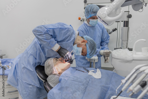 A dentist and an assistant surgeon perform an operation on patients to install dental implants. An elderly woman in a room with a microscope under the influence of anesthesia.