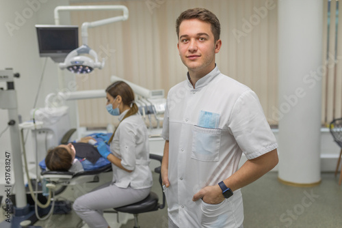 A male dentist looks at the camera against the background of a dental cabinet with a patient. Dental care and treatment concept