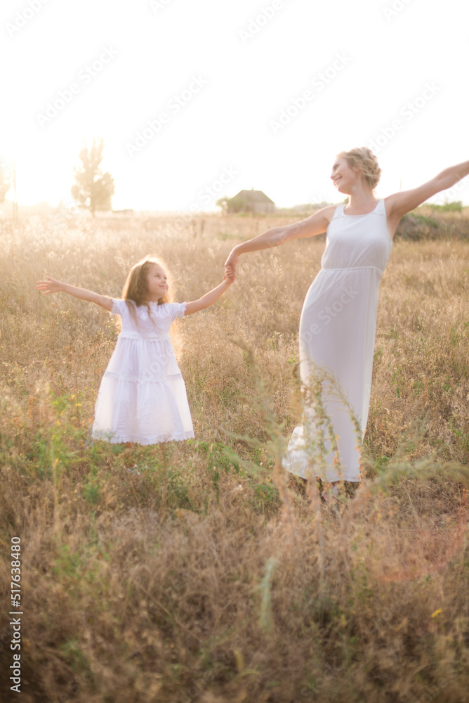 A cute little girl with long blond curly hair and her mother in a white summer dress and a straw boater hat in a field in the countryside in summer at sunset. Nature and Ecolife A cute little girl with long blond curly hair and her mother in a white summer dress and a straw boater hat in a field in the countryside in summer at sunset. Nature and Ecolife