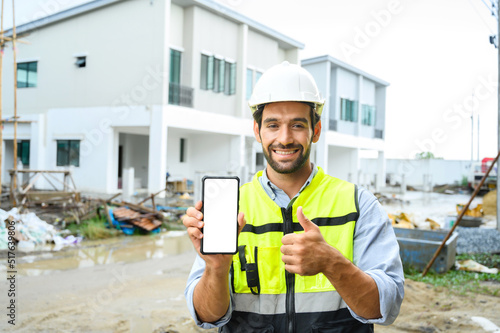 Young construction man smiling bearded in vest with white helmet demonstrating blank smartphone and showing thumb up to the camera while standing on building construction site. White screen, mockup.