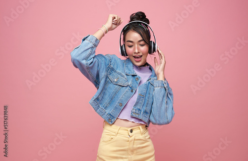 Happy beautiful Asian teen woman in yellow glasses listening music in headphones and dance on pink background.