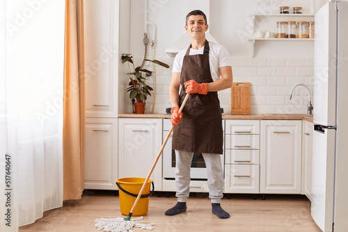 Indoor shot of smiling positive female wearing brown apron and rubber gloves, holding mop, washing floor in kitchen, looking at camera, being in good mood while doing household chores.