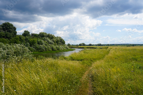 Narrow path through meadow by the river and sky with dramatic clouds on summer countryside landscape