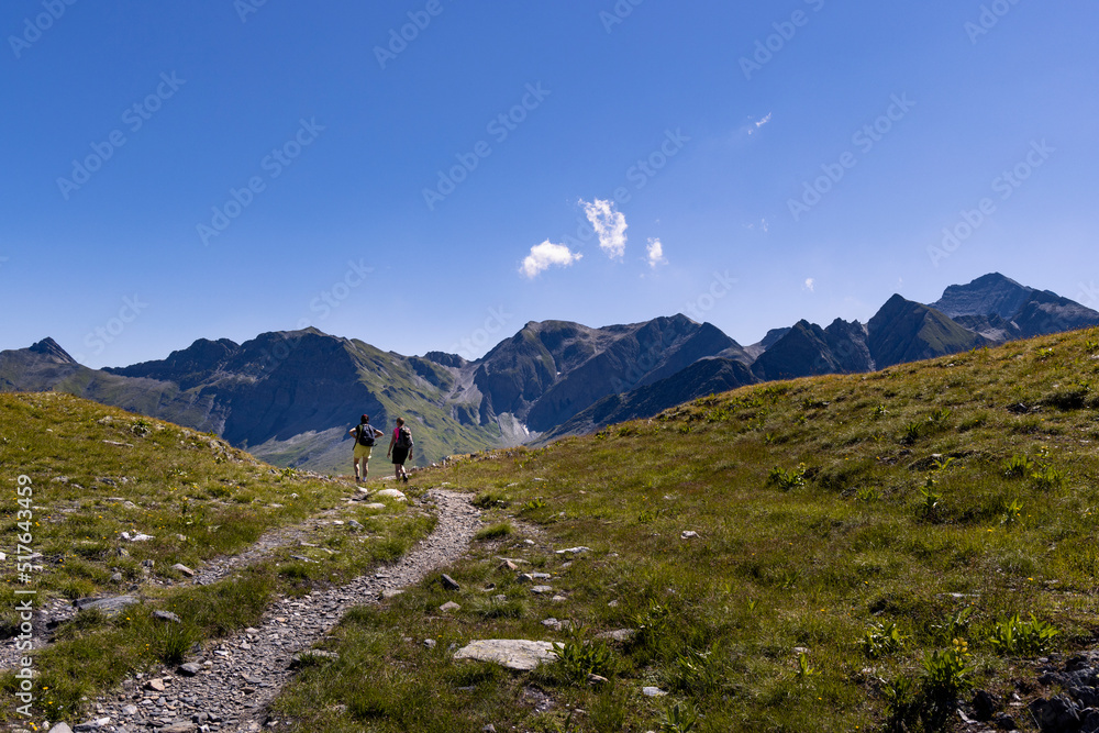 Two women walk on a winding alpine trail between mountains in late summer on a sunny day in the Greina Plateau, in Switzerland.