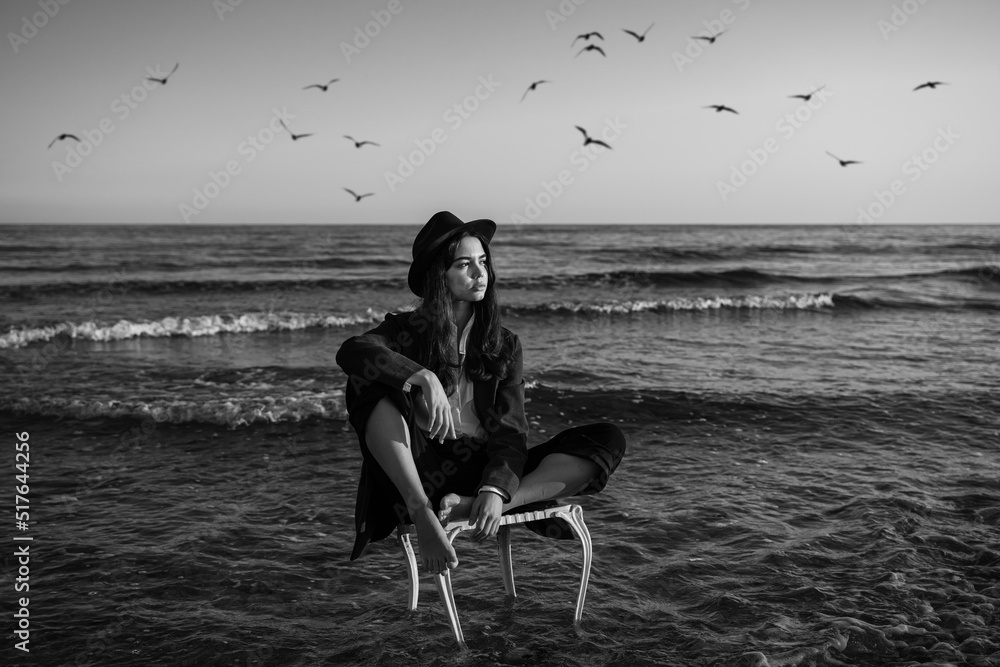 dreamer enjoys freedom by the sea. loneliness by the sea. girl in a formal elegant stylish suit, shirt and hat sitting on chair in waves of the sea.  seagulls in the sky. black and white horizontal 
