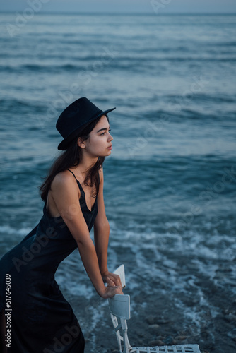 feelings and memories concept. girl in a hat near the sea leans on the back of a chair. Stylish feminine pensive slim girl looks at the camera. Blue sunset time sea composition. hat and dress. 