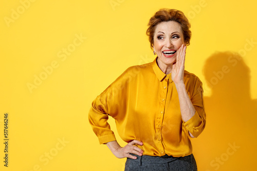 Smiling elderly woman shares her secrets with someone. Photo of attractive woman in yellow shirt on yellow background