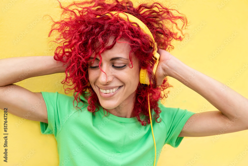 a happy latin woman with red afro hair with her eyes closed wearing a green t-shirt enjoys listening to music holding the yellow headphones with both hands.