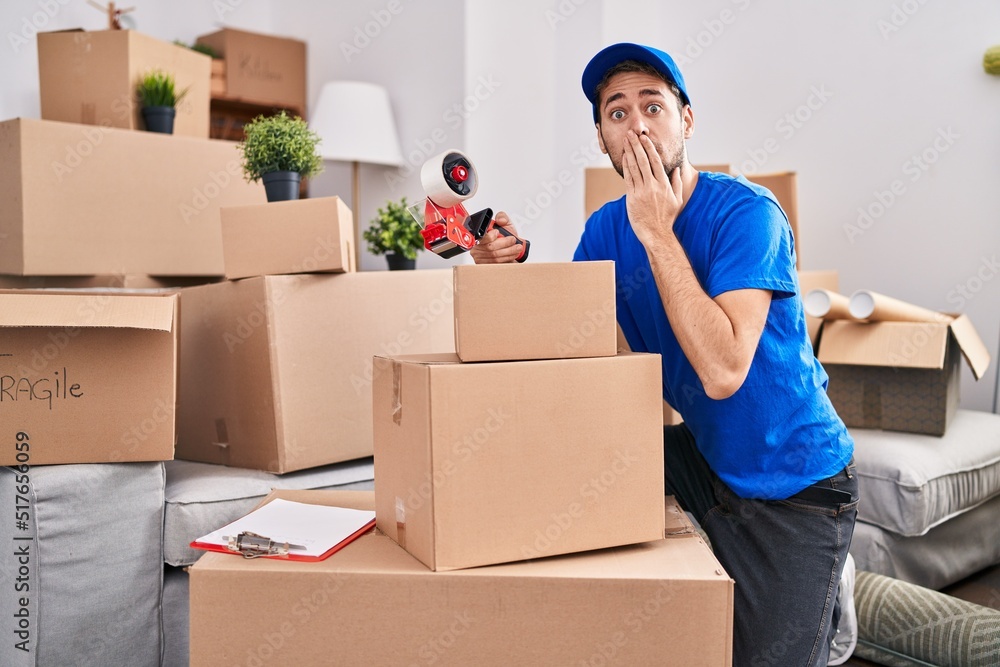 Hispanic man with beard working moving boxes covering mouth with hand, shocked and afraid for mistake. surprised expression Hispanic man with beard working moving boxes covering mouth with hand, shocked and afraid for mistake. surprised expression