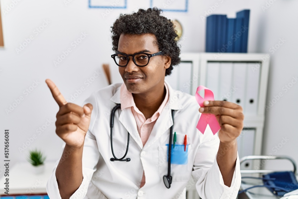 African doctor man holding pink cancer ribbon smiling happy pointing with hand and finger to the side African doctor man holding pink cancer ribbon smiling happy pointing with hand and finger to the side