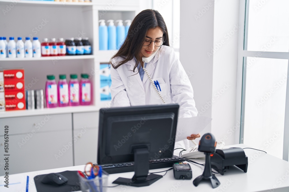 Young hispanic girl pharmacist talking by telephone reading document at pharmacy Young hispanic girl pharmacist talking by telephone reading document at pharmacy