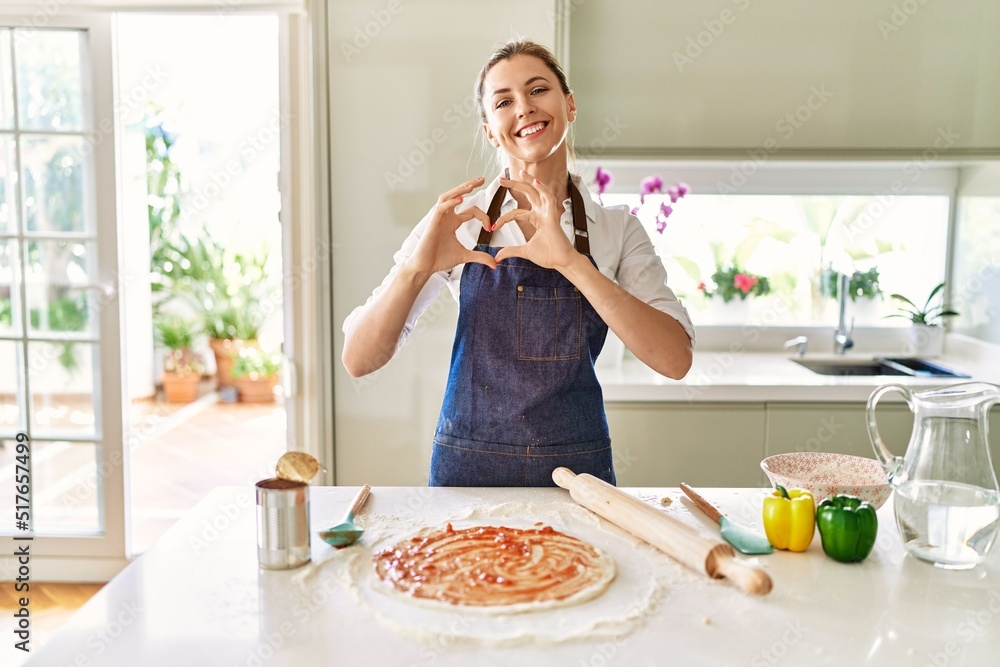 Beautiful blonde woman wearing apron cooking pizza smiling in love showing heart symbol and shape with hands. romantic concept.