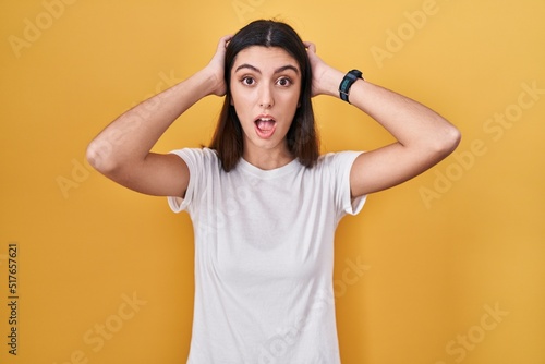 Young beautiful woman standing over yellow background crazy and scared with hands on head, afraid and surprised of shock with open mouth