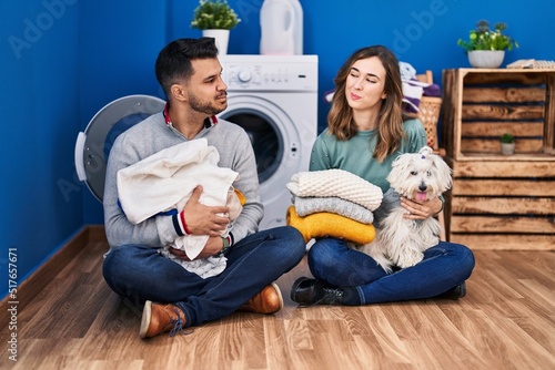 Young hispanic couple doing laundry sitting on the floor smiling looking to the side and staring away thinking.