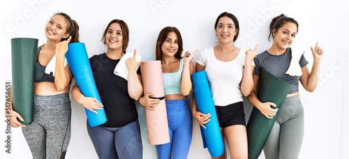 Group of women holding yoga mat standing over isolated background cheerful with a smile on face pointing with hand and finger up to the side with happy and natural expression