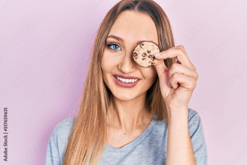 Young blonde girl holding cookie over eye looking positive and happy standing and smiling with a confident smile showing teeth Young blonde girl holding cookie over eye looking positive and happy standing and smiling with a confident smile showing teeth