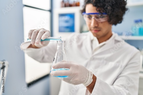 Young hispanic man wearing scientist uniform pouring liquid on test tube at laboratory