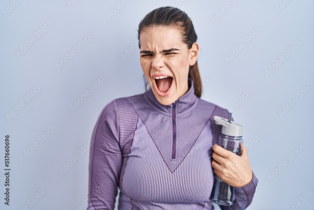 Beautiful woman wearing sportswear holding water bottle angry and mad screaming frustrated and furious, shouting with anger. rage and aggressive concept. Beautiful woman wearing sportswear holding water bottle angry and mad screaming frustrated and furious, shouting with anger. rage and aggressive concept.