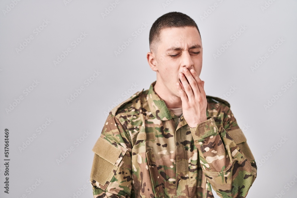 Young man wearing camouflage army uniform bored yawning tired covering mouth with hand. restless and sleepiness.