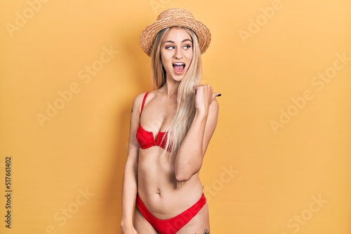 Young caucasian woman wearing bikini and summer hat smiling with happy face looking and pointing to the side with thumb up.