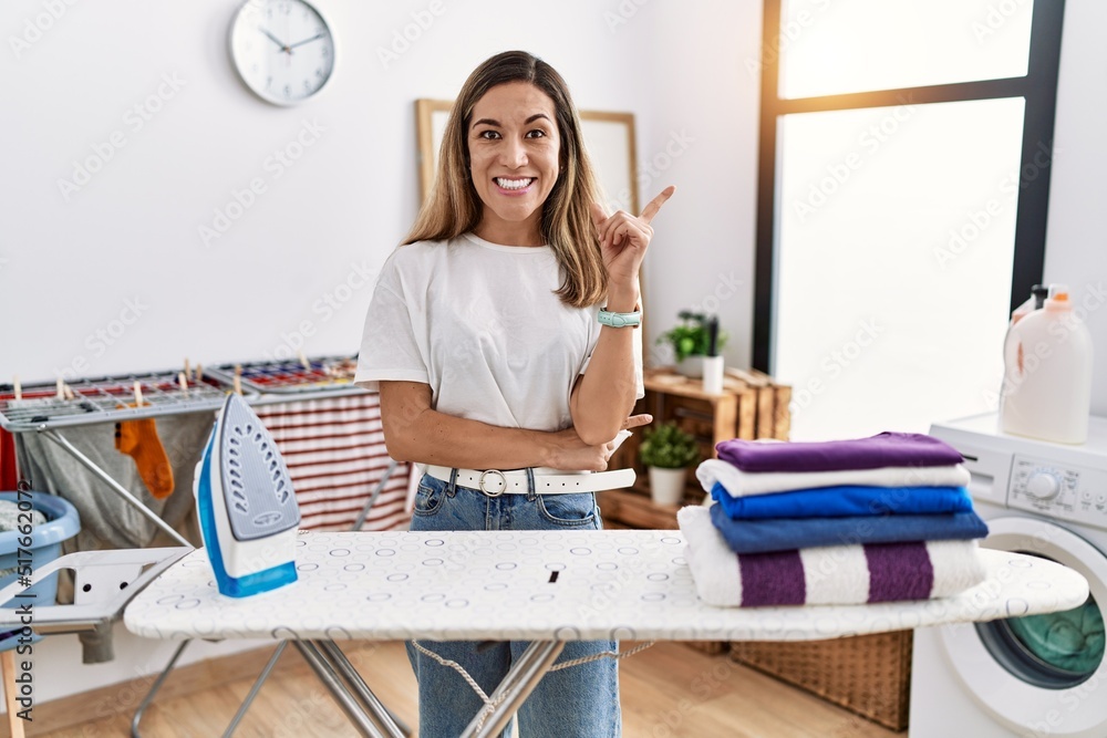 Young hispanic woman ironing clothes at laundry room with a big smile on face, pointing with hand and finger to the side looking at the camera. Young hispanic woman ironing clothes at laundry room with a big smile on face, pointing with hand and finger to the side looking at the camera.