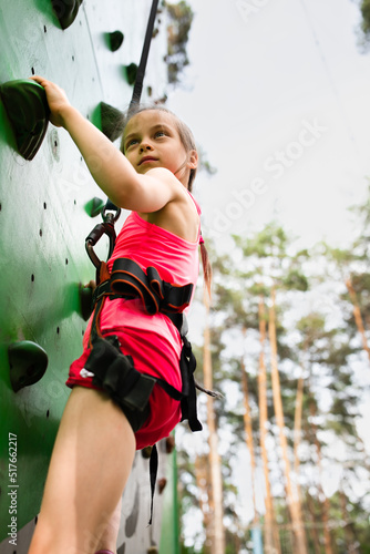 Little adorable girl in harness climbing on rock climbing wall in sky entertainment park in summer