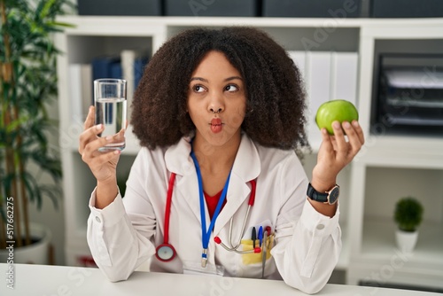 Young african american dietitian woman holding fresh apple and water making fish face with mouth and squinting eyes, crazy and comical.