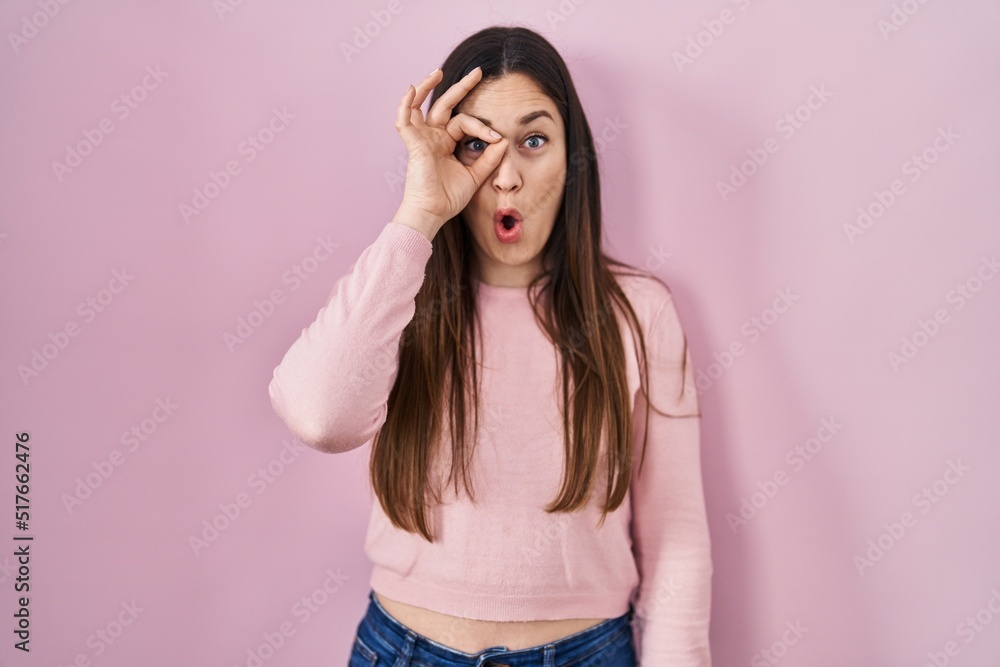 Young brunette woman standing over pink background doing ok gesture shocked with surprised face, eye looking through fingers. unbelieving expression. Young brunette woman standing over pink background doing ok gesture shocked with surprised face, eye looking through fingers. unbelieving expression.