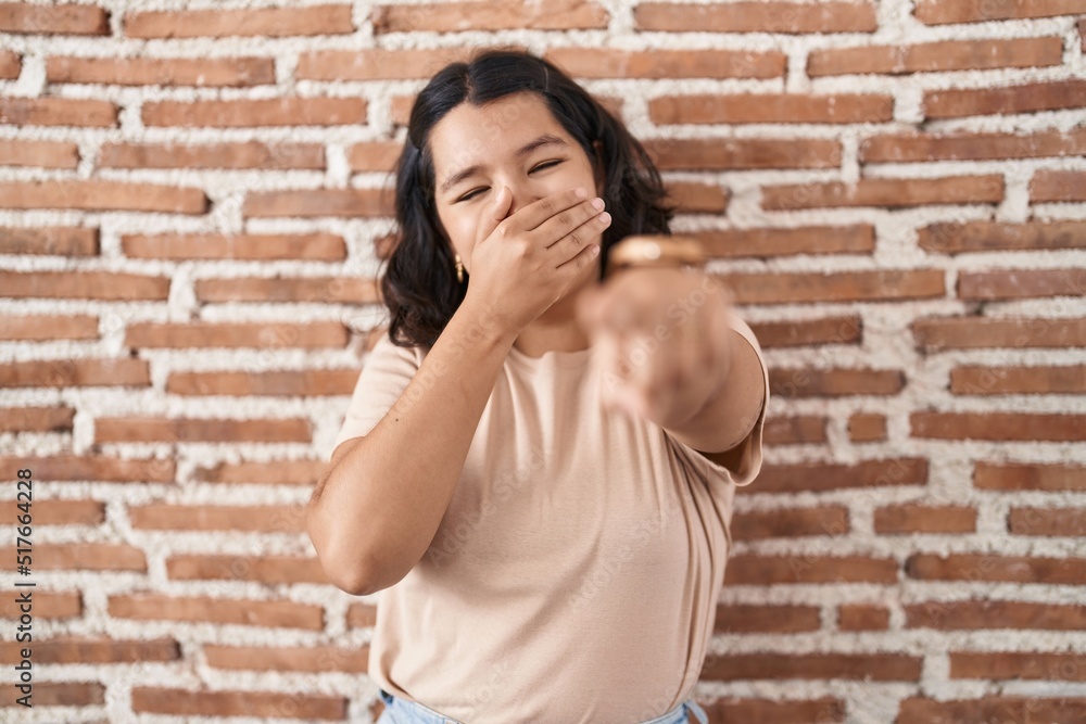 Young hispanic woman standing over bricks wall laughing at you, pointing finger to the camera with hand over mouth, shame expression Young hispanic woman standing over bricks wall laughing at you, pointing finger to the camera with hand over mouth, shame expression