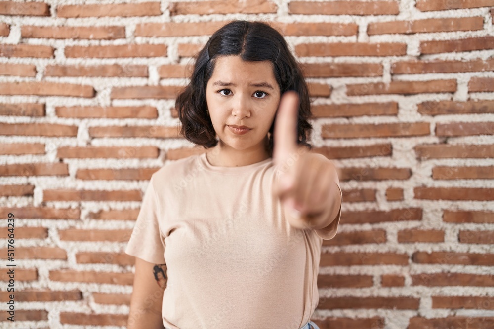 Young hispanic woman standing over bricks wall pointing with finger up and angry expression, showing no gesture Young hispanic woman standing over bricks wall pointing with finger up and angry expression, showing no gesture