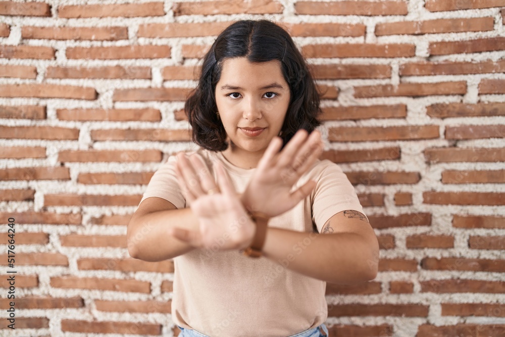 Young hispanic woman standing over bricks wall rejection expression crossing arms and palms doing negative sign, angry face Young hispanic woman standing over bricks wall rejection expression crossing arms and palms doing negative sign, angry face