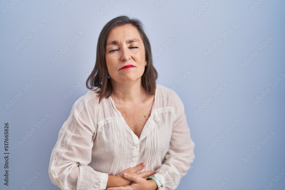 Middle age hispanic woman standing over blue background with hand on stomach because indigestion, painful illness feeling unwell. ache concept. Middle age hispanic woman standing over blue background with hand on stomach because indigestion, painful illness feeling unwell. ache concept.