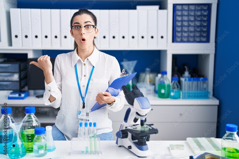 Young brunette woman working at scientist laboratory surprised pointing with hand finger to the side, open mouth amazed expression.