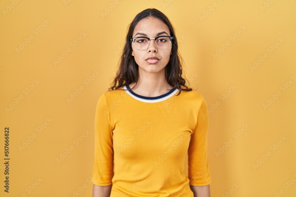 Obraz premium Young brazilian woman wearing glasses over yellow background with serious expression on face. simple and natural looking at the camera.