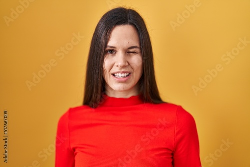 Young hispanic woman standing over yellow background winking looking at the camera with sexy expression, cheerful and happy face.