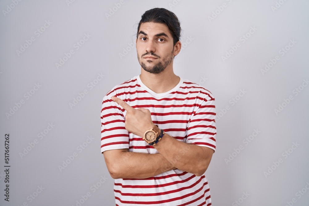 Hispanic man with long hair standing over isolated background pointing with hand finger to the side showing advertisement, serious and calm face Hispanic man with long hair standing over isolated background pointing with hand finger to the side showing advertisement, serious and calm face