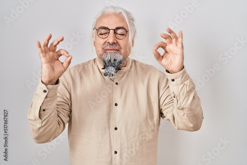 Middle age man with grey hair standing over isolated background relax and smiling with eyes closed doing meditation gesture with fingers. yoga concept.