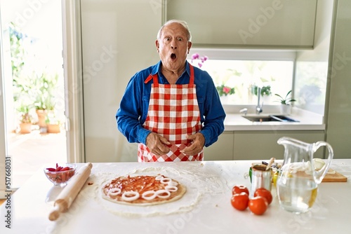 Senior man with grey hair cooking pizza at home kitchen scared and amazed with open mouth for surprise, disbelief face