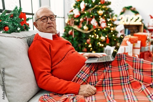 Obraz na plátně Senior man with grey hair using laptop sitting by christmas tree thinking attitu