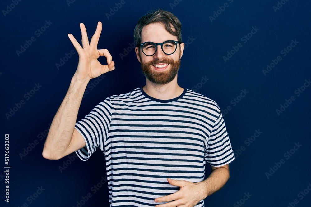 Caucasian man with beard wearing striped t shirt and glasses smiling positive doing ok sign with hand and fingers. successful expression.
