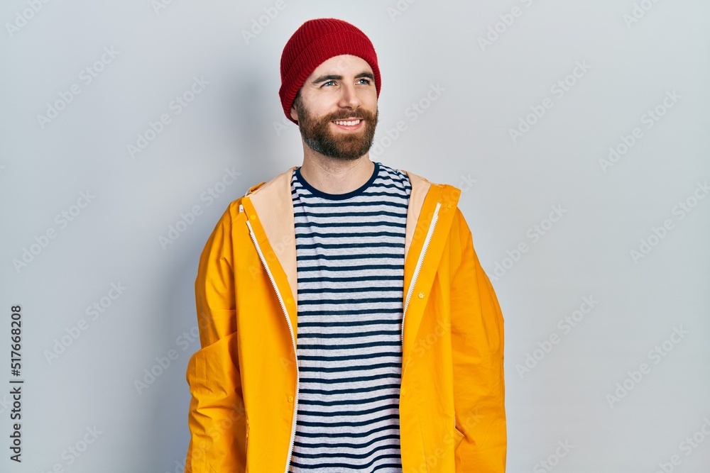 Caucasian man with beard wearing yellow raincoat looking away to side with smile on face, natural expression. laughing confident.