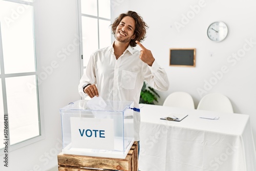 Young hispanic man voting putting envelop in ballot box smiling cheerful showing and pointing with fingers teeth and mouth. dental health concept. photo