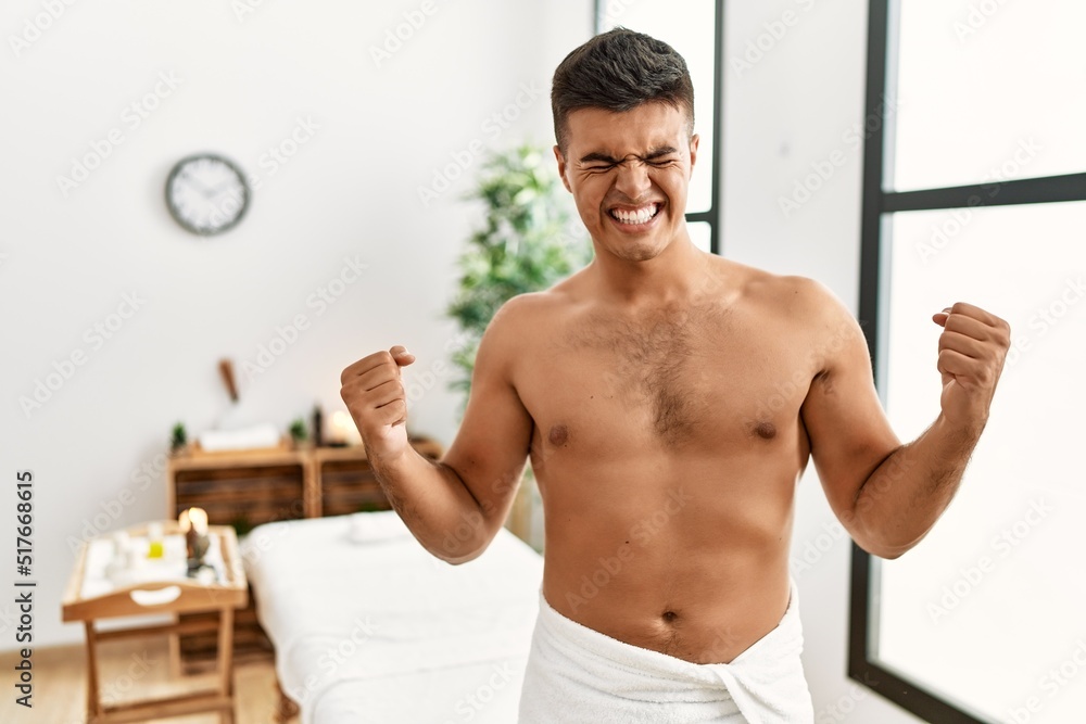 Obraz premium Young hispanic man standing shirtless at spa center very happy and excited doing winner gesture with arms raised, smiling and screaming for success. celebration concept.