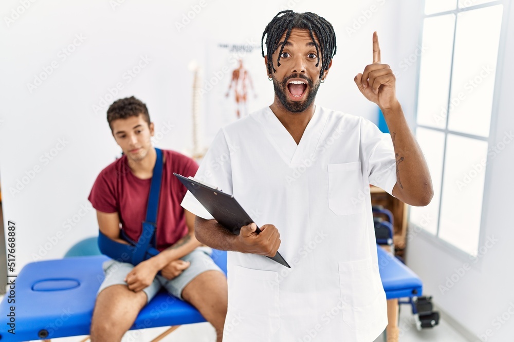 Obraz premium Young hispanic man working at pain recovery clinic with a man with broken arm pointing finger up with successful idea. exited and happy. number one.