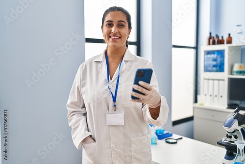 Young hispanic woman wearing scientist uniform using smartphone at laboratory