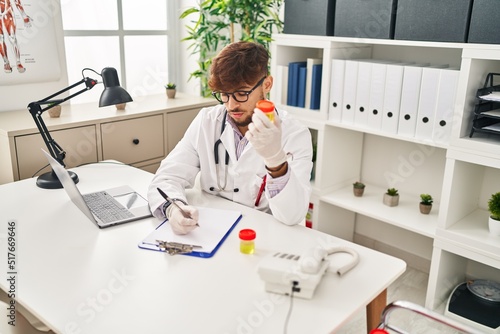 Young arab man wearing doctor uniform writing on document analysing urine test tube at clinic